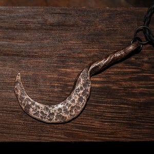 May include: A rustic, metal pendant necklace featuring a curved, hook-shaped charm. The charm has a textured, aged appearance with a dark, braided leather cord. The pendant is a silver color and rests on a dark wooden surface.