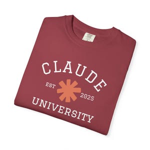 May include: A maroon t-shirt featuring the words "CLAUDE UNIVERSITY" in white. The text is arched above an orange starburst design, with "EST 2025" below "CLAUDE".