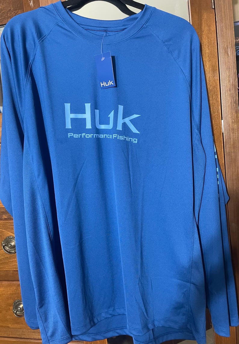 May include: A long-sleeved, blue performance fishing shirt. The shirt features the brand name "Huk" in large, light blue letters across the chest, with "Performance Fishing" below. A small tag with the "Huk" logo hangs from the collar.
