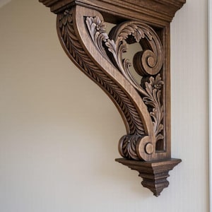 Puede incluir: Un ménsula de madera tallada de color marrón oscuro con un diseño decorativo de flores y hojas. La ménsula está montada en una pared de color claro, mostrando sus detalles ornamentados y su estilo arquitectónico. Es un elemento decorativo.