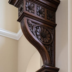 Könnte beinhalten: Ein dunkelbrauner, verzierter Wandhalter aus Holz mit floralen Schnitzereien. Der Halter hat eine rechteckige Oberseite mit geschnitzten Blumen, eine geschwungene Stütze mit einem großen Blumenmedaillon und einen dekorativen Sockel.