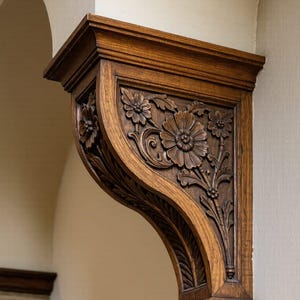 Könnte beinhalten: Ein geschnitztes Holzkonsole mit floralen Details. Die dunkelbraune Konsole ist an einer hellen Wand befestigt. Das florale Design umfasst große, detaillierte Blumen und Blätter. Es ist ein dekoratives architektonisches Element.