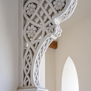 Puede incluir: Un soporte arquitectónico de madera blanca y ornamentada con diseños florales y entrelazados. El soporte está montado en una pared blanca, con una ventana arqueada visible en el fondo. Las intrincadas tallas del soporte añaden un elemento decorativo.