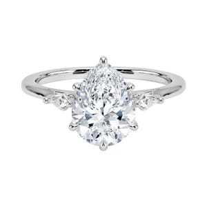 May include: A silver engagement ring featuring a large, pear-shaped diamond as the centerpiece. Two smaller diamonds flank the main stone. The ring has a polished, reflective surface, and the diamonds sparkle brightly against a white background.