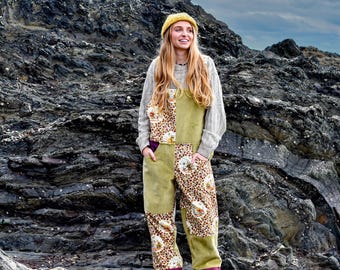 May include: A woman wearing a yellow beanie, gray sweater, and olive green overalls with leopard print patches. The overalls have purple accents and are paired with dark shoes. The background features a rocky landscape under a cloudy sky.