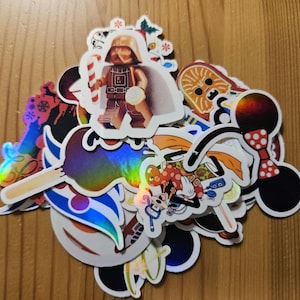 Könnte beinhalten: Eine Sammlung bunter Aufkleber mit verschiedenen Charakteren und Designs. Enthält eine Darth-Vader-Figur mit einer Zuckerstange, einen Chewbacca-Aufkleber und Mickey- und Minnie-Mouse-Aufkleber. Einige Aufkleber haben einen holografischen Effekt.