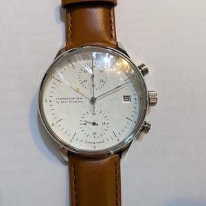 Może przedstawiać: Srebrny zegarek chronograf z białą tarczą i brązowym skórzanym paskiem. Na tarczy zegarka widnieje napis "CHRONOGRAPH 1844 by Skov Andersen". Zegarek posiada trzy sub-tarcze i okienko datownika.