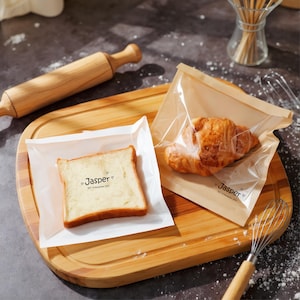 May include: A wooden chopping board displays a slice of bread and a croissant, each in a clear bag. The bags are labelled "Jasper" with a heart symbol. A rolling pin and whisk are in the background.