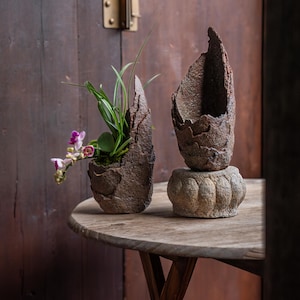 May include: Two textured, brown, organic-shaped planters on a round wooden table. One planter holds a small plant with green leaves and purple flowers. The other planter sits atop a decorative stone base. The table has a dark wood finish.
