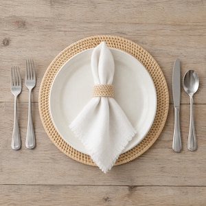 May include: A white ceramic dinner plate sits on a round woven rattan placemat. A white linen napkin with frayed edges is folded and secured with a matching woven rattan napkin ring. Two silver forks are on the left, and a knife and spoon are on the right.