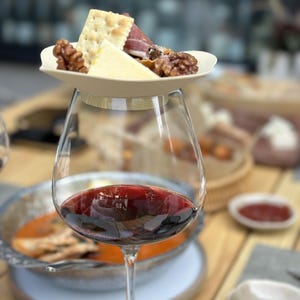 Puede incluir: Una copa de vino tinto con un plato pequeño con galletas, queso, nueces y prosciutto encima. El fondo muestra un cuenco de comida y otros elementos de comedor.
