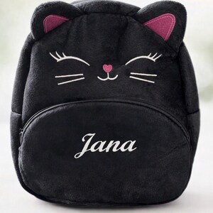 May include: A plush black backpack shaped like a cat head with pink inner ears, embroidered white eyes, whiskers, and a pink nose. The front pocket features the name Jana embroidered in white script. The bag has a soft, fuzzy texture throughout.