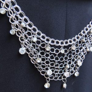 May include: Silver-toned statement necklace with a chainmail design. The necklace features multiple strands of interconnected links, adorned with dangling clear, round rhinestones. The jewelry is displayed against a dark background.