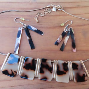 May include: A jewelry set consisting of a necklace and two earrings. The necklace features five rectangular, tortoise-shell patterned acrylic pieces separated by gold-tone metal bars on a snake chain. The earrings have three dangling rectangular pieces.
