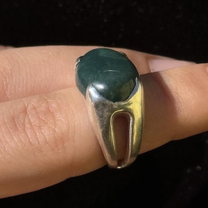 Könnte beinhalten: Ein Silberring mit einem großen, ovalen, dunkelgrünen Edelstein. Der Ring hat ein geteiltes Schaftdesign, und der Edelstein ist in einer Lünettenfassung gefasst. Der Ring wird an einem Finger gezeigt.