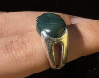 May include: A silver ring featuring a large, oval-shaped, dark green gemstone. The ring has a split shank design, and the gemstone is set in a bezel setting. The ring is shown on a finger.