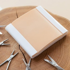 May include: A light tan, rectangular practice pad with white edges is surrounded by several stainless steel surgical instruments, including scissors and forceps. The items are arranged on a round, wooden surface with a natural grain.