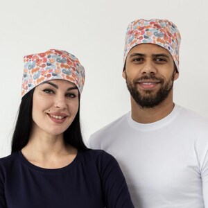 May include: Two people wearing matching floral print scrub caps and long-sleeved shirts. One person wears a navy blue shirt, the other a white shirt. Both are smiling, posing in front of a white background.