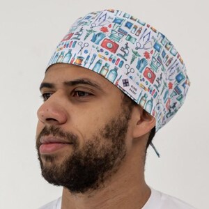 May include: A white surgical scrub cap featuring a repeating pattern of medical instruments in blue, red, and green. The cap is tied at the back and worn by a person in a white long-sleeved top.