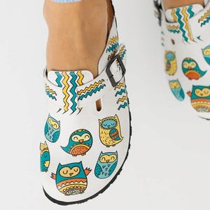 May include: White clogs with a black sole and buckle fastening. The clogs feature colourful owl illustrations and zig-zag patterns in yellow, teal, and blue. These clogs are designed for comfort and style.