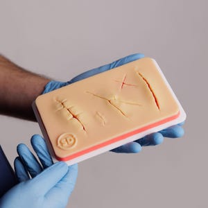 Könnte beinhalten: Ein hautfarbenes medizinisches Trainingspad mit simulierten Rissen und Nähten. Das Pad hat einen weißen Rand mit rotem Akzent. Das Pad wird von einer Person mit blauen Handschuhen gehalten. Das Pad hat die Buchstaben "SD" und ein kreisförmiges Logo.