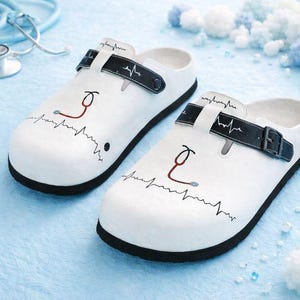 May include: White clogs with black soles and straps, featuring a stethoscope and EKG design. The medical-themed shoes have a buckle closure and are decorated with a red stethoscope graphic and a black heartbeat line.