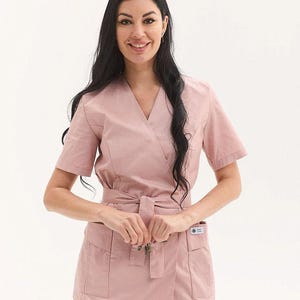 Puede incluir: Una blusa de uniforme rosa claro estilo envolvente con mangas cortas, cinturón anudado y dos bolsillos delanteros. La blusa de uniforme está hecha de una tela suave y la lleva una persona con pelo largo y negro.