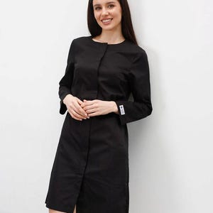 Puede incluir: Vestido negro de manga larga con cuello mandarín y dobladillo a la rodilla. El vestido tiene cierre de botones y una pequeña etiqueta en la manga izquierda. La modelo sonríe, sobre fondo blanco.