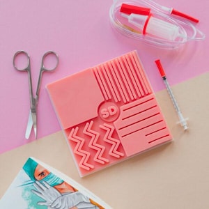 Könnte beinhalten: Ein rosa medizinisches Trainingspad mit verschiedenen strukturierten Mustern, einer Schere, einer Spritze und einem medizinischen Gerät mit Schläuchen. Das Pad hat die Buchstaben "SD" in einem Kreis. Der Hintergrund ist rosa und beige.