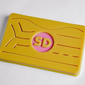 Könnte beinhalten: Ein rechteckiges, gelbes Objekt mit einem rosa Kreis in der Mitte, der die gelben Buchstaben "SD" trägt. Das Objekt hat eine Reihe horizontaler Linien und ein einzigartiges Ausschnittdesign. Die Kanten sind rosa und weiß umrandet.