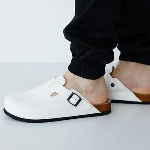 May include: White leather clogs with a black sole and a buckle closure. The clogs have a cork footbed and are worn with black pants. The shoes are a slip-on style.