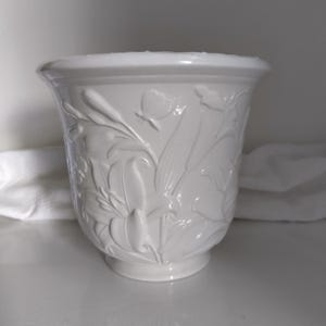 May include: A white ceramic planter featuring a raised relief design of lilies and a butterfly. The vessel has a wide, flared rim, a tapered body, and a circular base. The surface is smooth and glossy, with the floral motifs integrated into the structure.