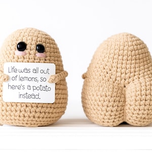 Puede incluir: Dos figuras de crochet con forma de patata. Una sostiene un cartel que dice: "Life was all out of lemons, so here's a potato instead". Las patatas son de color beige con ojos negros y mejillas rosas. La otra patata está de espaldas.