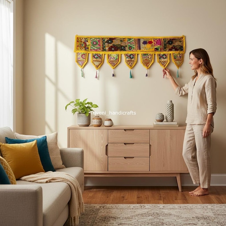May include: A vibrant, yellow and multi-colored textile wall hanging with a patchwork design. The decorative piece features intricate embroidery and hanging tassels. The wall hanging is displayed above a wooden cabinet in a living room setting.