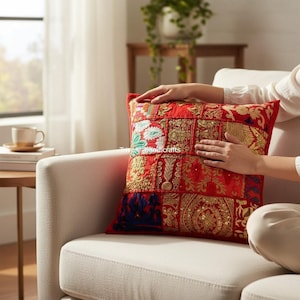 Op de afbeelding: Een rood decoratief kussen met een patchwork ontwerp. Het kussen heeft gouden borduursels, pailletten en diverse stofpatronen. Het vierkante kussen ligt op een witte bank.