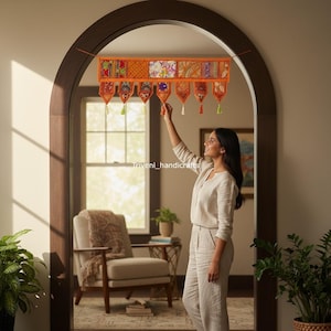 May include: An orange and multi-colored fabric door hanging with a patchwork design. The hanging has a series of decorative pendants with tassels. The image shows a woman adjusting the hanging in a doorway.