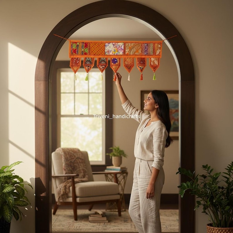May include: An orange and multi-colored fabric door hanging with a patchwork design. The hanging has a series of decorative pendants with tassels. The image shows a woman adjusting the hanging in a doorway.