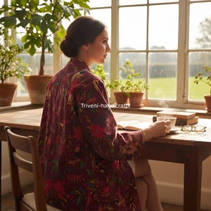 May include: A burgundy floral print robe with long sleeves. The robe is worn by a person sitting at a wooden table, holding a cup of tea. The text "Triveni-handicrafts" is visible on the robe.