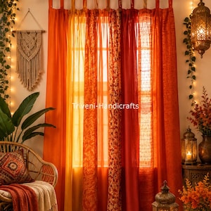 May include: A set of orange and red curtains with tab tops hanging on a black rod, featuring a mix of solid and patterned fabric panels. The text Triveni-Handicrafts is visible in the center. The curtains are illuminated by warm string lights.
