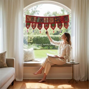 May include: A red and multi-colored textile wall hanging with intricate patterns and tassels. The banner is suspended in front of a large window with white curtains. The text "Triveni_handicrafts" is visible.