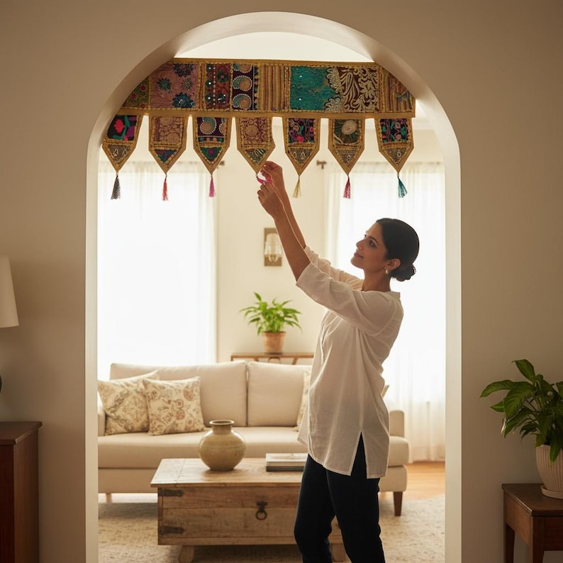May include: A woman is shown adjusting a decorative archway valance. The valance is made of colorful, patterned fabric panels with hanging tassels. The archway is in a living room with a sofa, coffee table, and plants.