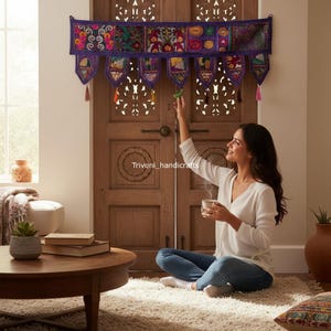 May include: A decorative purple wall hanging with colorful embroidered panels and tassels. The wall hanging is above a wooden door. A woman is sitting on the floor, holding a cup.