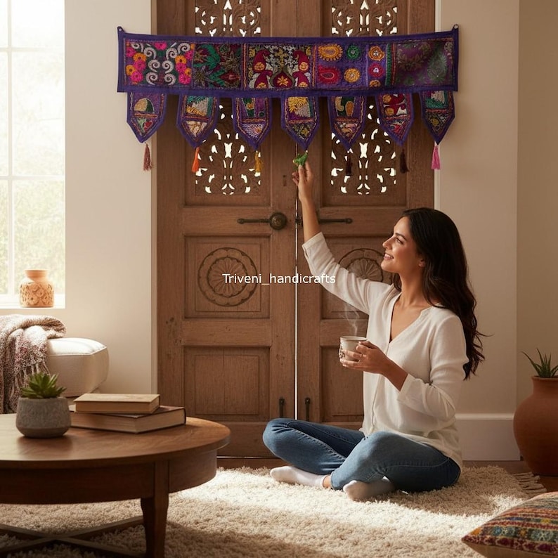 May include: A decorative purple wall hanging with colorful embroidered panels and tassels. The wall hanging is above a wooden door. A woman is sitting on the floor, holding a cup.