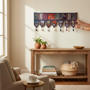 May include: A decorative wall hanging with a dark blue border and colorful patterned panels. Each panel has a tassel. The wall hanging is displayed above a wooden console table with plants, books, and a woven basket.