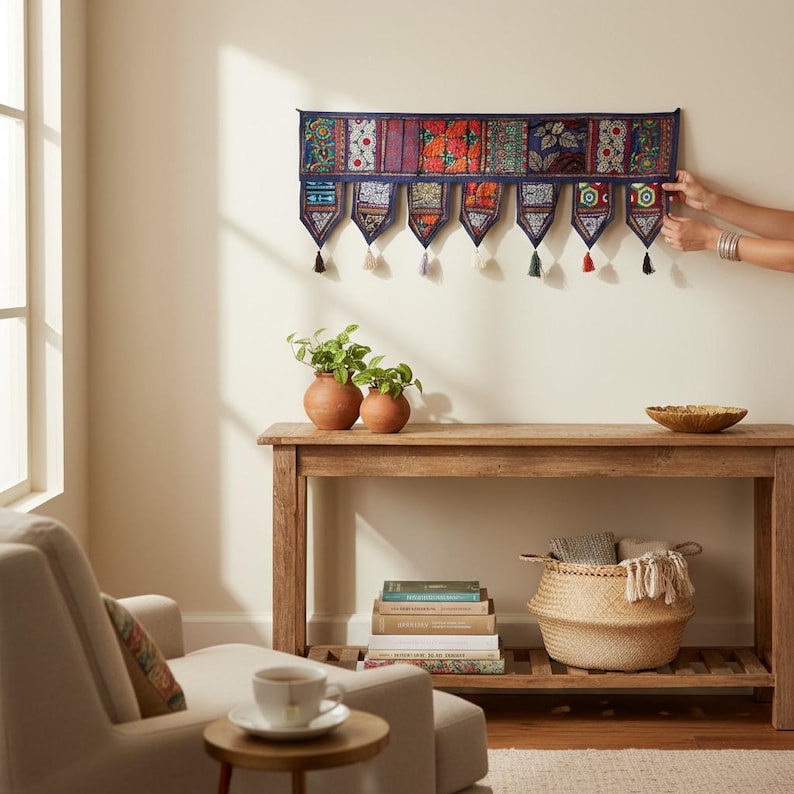 May include: A decorative wall hanging with a dark blue border and colorful patterned panels. Each panel has a tassel. The wall hanging is displayed above a wooden console table with plants, books, and a woven basket.