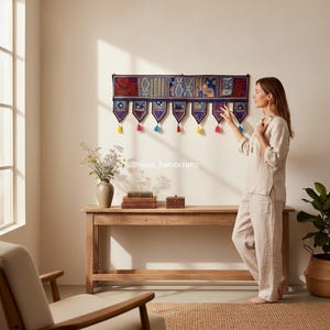 May include: A decorative wall hanging with a rectangular top and seven pendant-shaped bottoms. The top features a patchwork design in red, blue, and gold. Each pendant has a tassel in a different color. The text "Triveni_handicrafts" is visible.