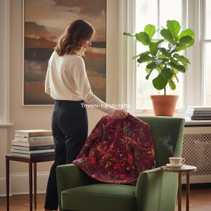 May include: A burgundy floral patterned jacket is draped over a green armchair. The jacket features a detailed design with various colors. A person is holding the jacket. The scene is set in a well-lit room with a plant and books.