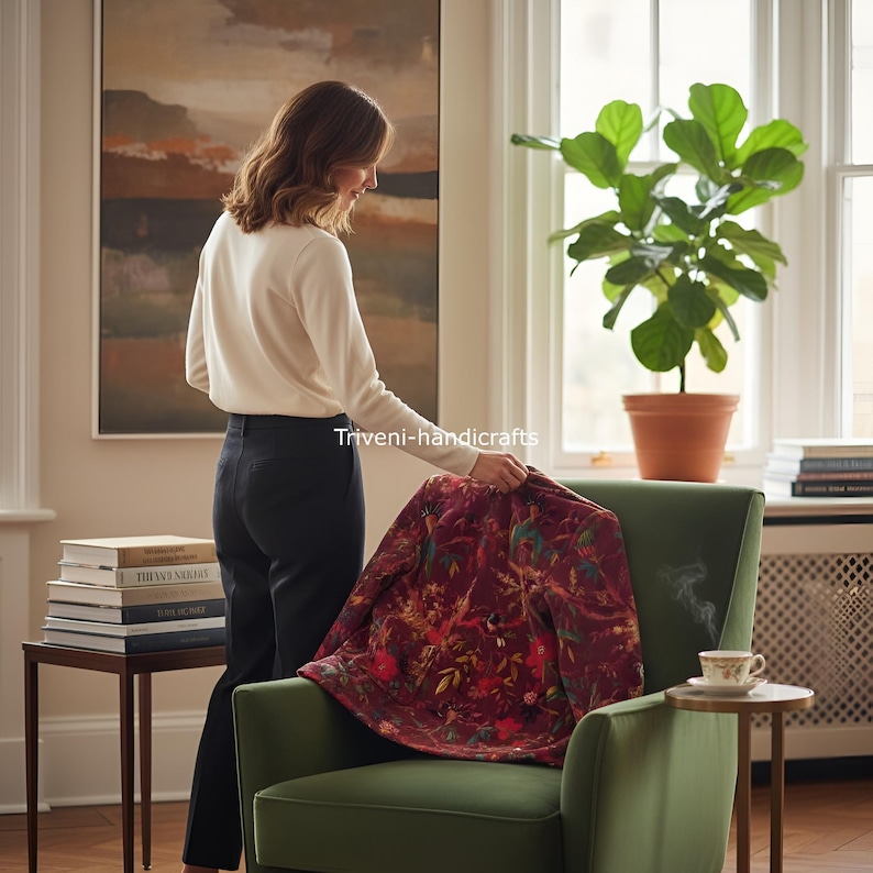 May include: A burgundy floral patterned jacket is draped over a green armchair. The jacket features a detailed design with various colors. A person is holding the jacket. The scene is set in a well-lit room with a plant and books.