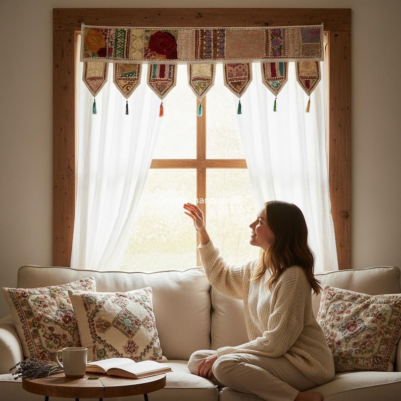 May include: A decorative window valance with a patchwork design in shades of beige, red, and blue, adorned with tassels. The valance hangs above white curtains, with a woman sitting on a cream-colored sofa.