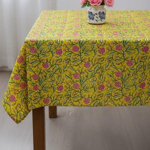 Puede incluir: Un mantel amarillo con un estampado floral de flores rosas y vides verdes cubre una mesa de madera. Un pequeño jarrón con rosas rosas está sobre la mesa. El mantel tiene un diseño brillante y alegre.